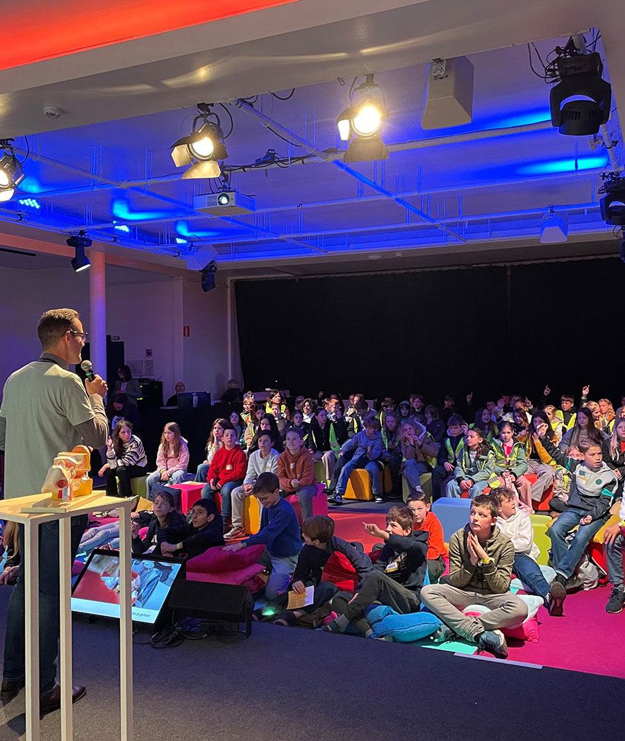 A speaker stands in front of a large group of children sitting in a dark room with colorful carpets and lighting.
